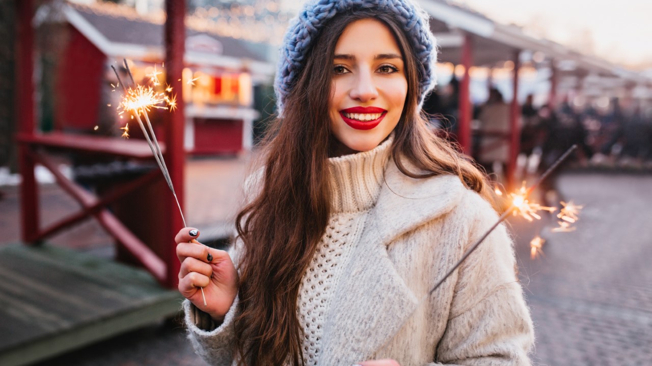 Portrait of graceful brown-haired woman in stylish white coat holding Bengal lights. Outdoor photo of romantic european girl in blue beret posing with sparklers on blur city background..